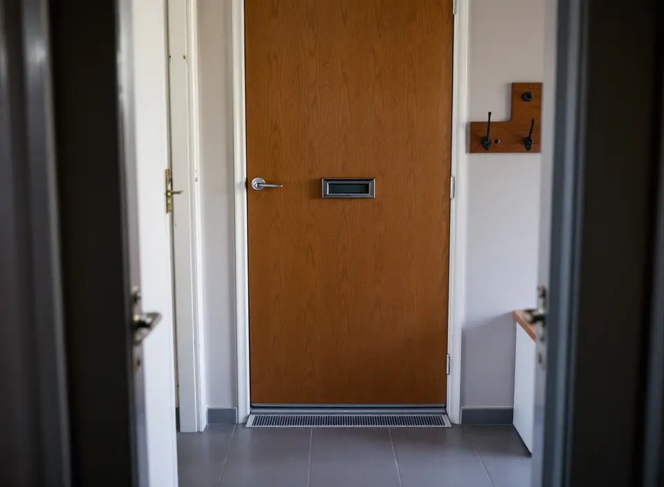 Een gang met een houten voordeur, met een metalen brievenbus op heuphoogte en een rooster onder de deur